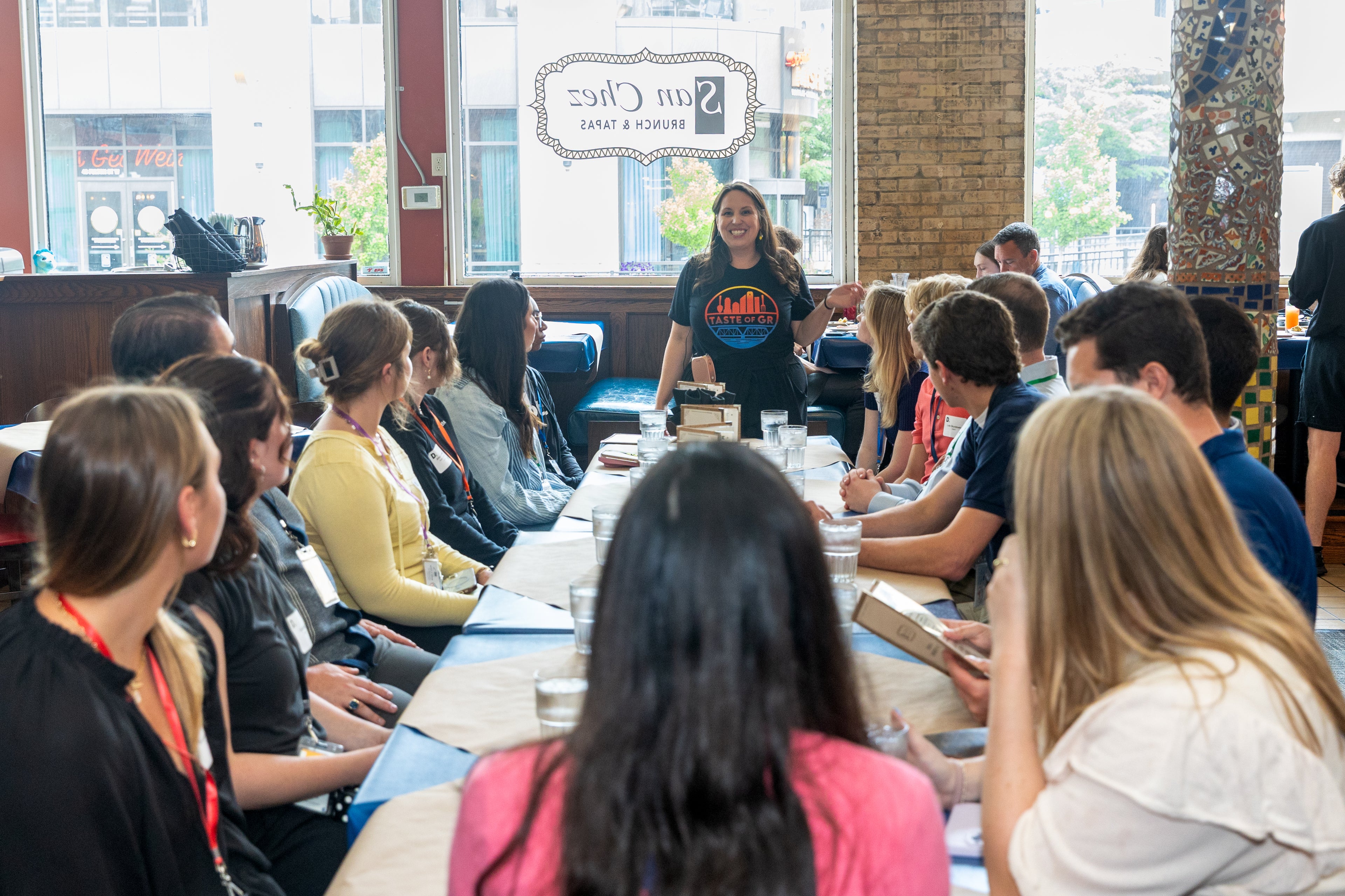 Group of people sitting at a long table, doing a food tour, with a woman tour guide standing and speaking.